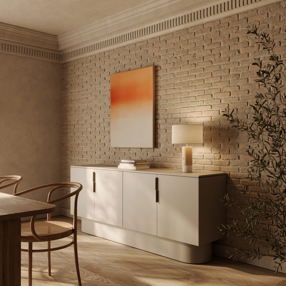 Sano Table Lamp lit on a white sideboard against a brick wall with orange artwork and bentwood chairs