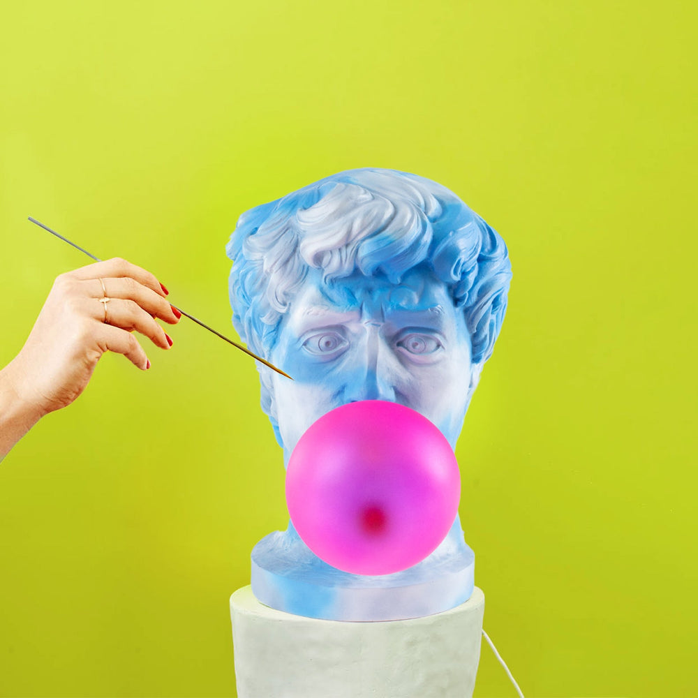 Seletti Wonder Cloud Bust Lamp, a hand about to pop the pink bubble with a pin against a lime backdrop