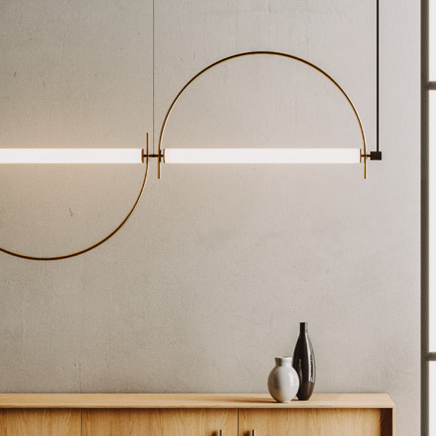 Two Arc Pendant Lamps in aged gold joined in an S-curve, lit, above a wooden sideboard with vases