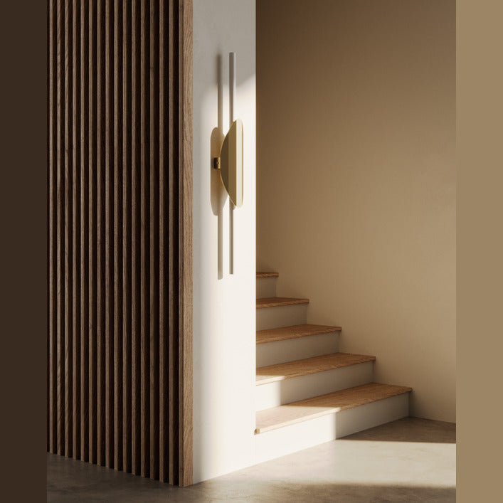 Xago Wall Lamp in gold lit beside a wooden staircase with a slatted wood screen