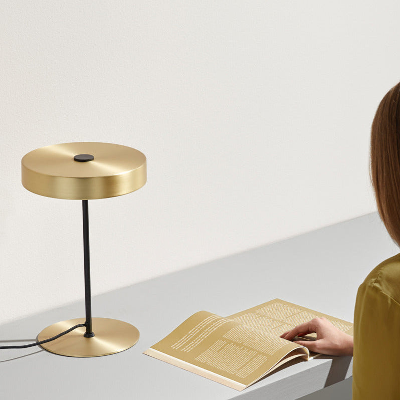 Ambor Table Lamp on a white desk beside a woman reading a magazine