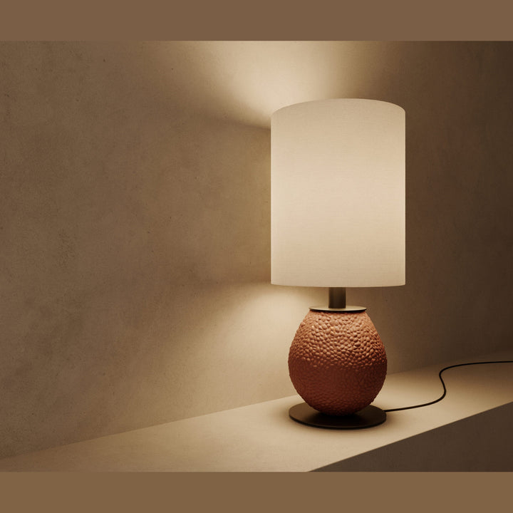 Anada Table Lamp in textured terracotta ceramic, lit, casting a soft glow on a plaster wall