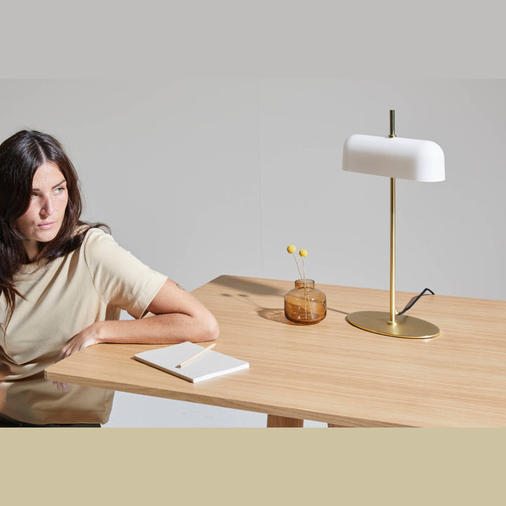 Atil Table Lamp in matt brass on an oak table beside an amber vase, a woman seated nearby
