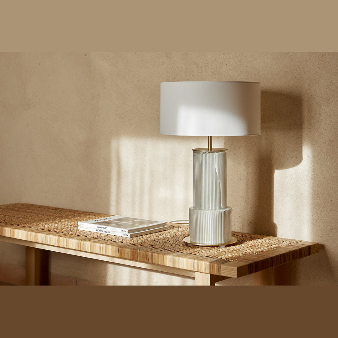 Atina Table Lamp with white drum shade on a woven rattan bench in warm sunlight against a plaster wall