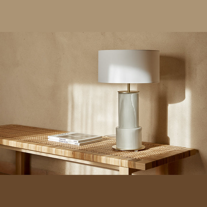 Atina Table Lamp with white drum shade on a woven rattan bench in warm sunlight against a plaster wall