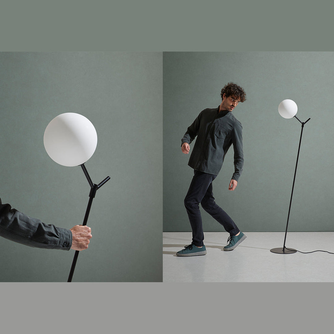 Atom Floor Lamp in black against a sage green wall with a man walking past, plus a hand holding the stem