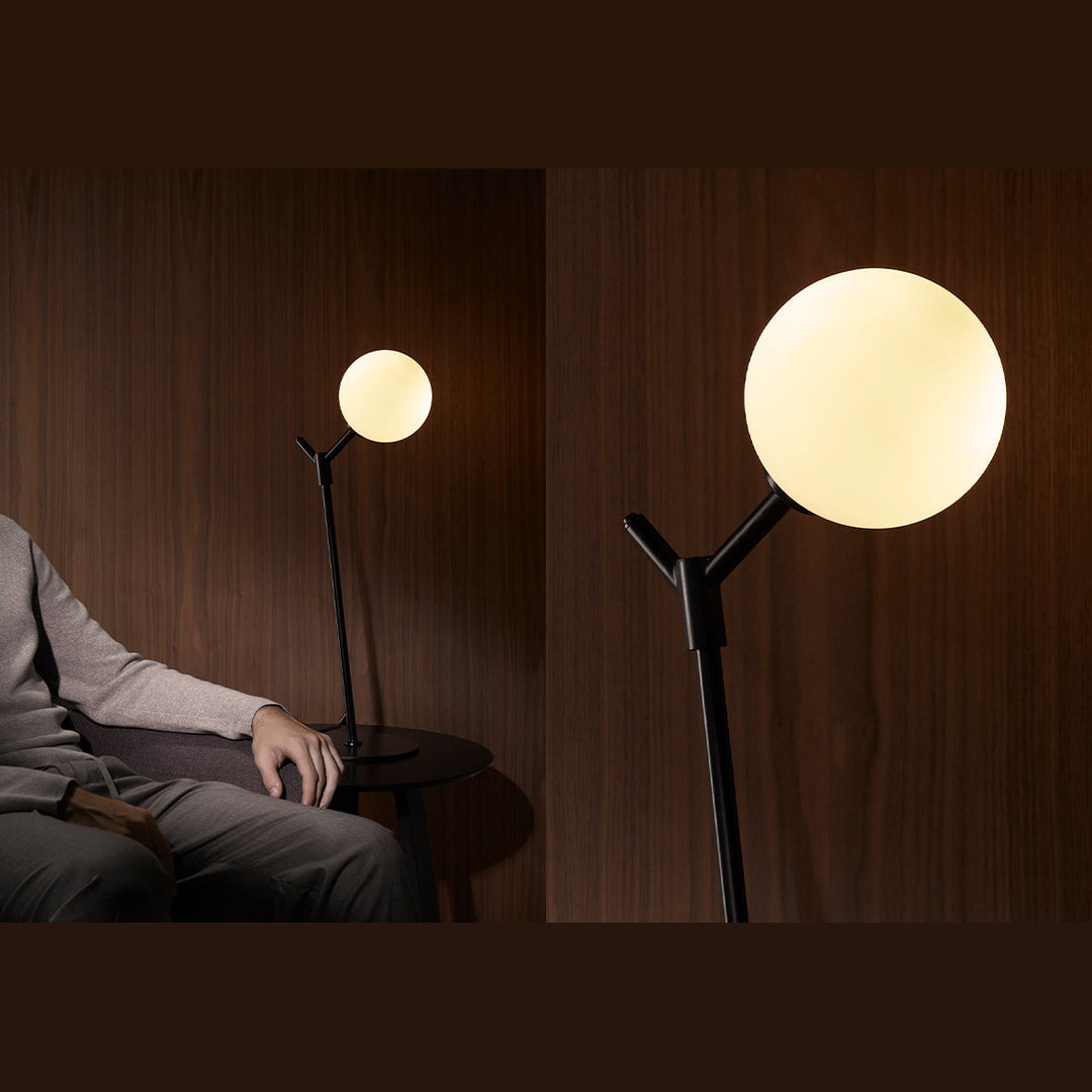 Atom Table Lamp in black, lit, on a side table beside a seated man against walnut panelling, plus a globe close-up