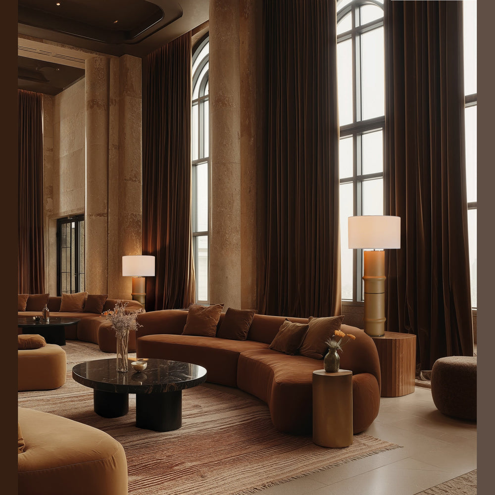 Bambu Floor Lamp in mustard, lit, beside a curved velvet sofa in a tall-windowed lounge with brown drapes