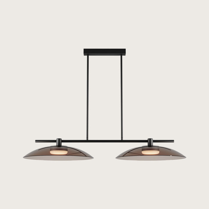 Betty Linear Pendant Linear Pendant, black bar with two wide smoked glass shades, lit, on a pale backdrop by Aromas del Campo