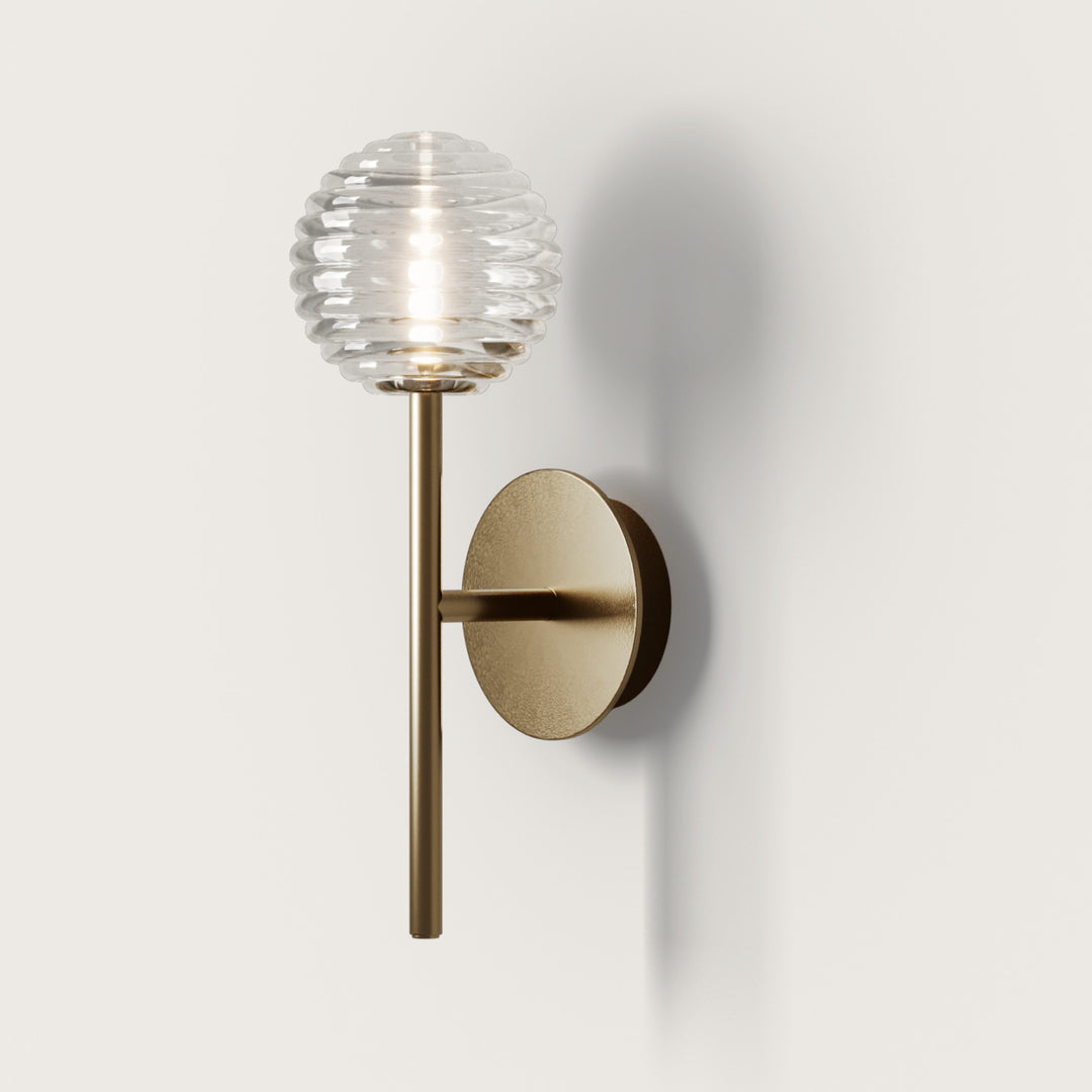 Doul Wall Lamp in aged gold with a ribbed clear glass globe, lit, by Aromas del Campo