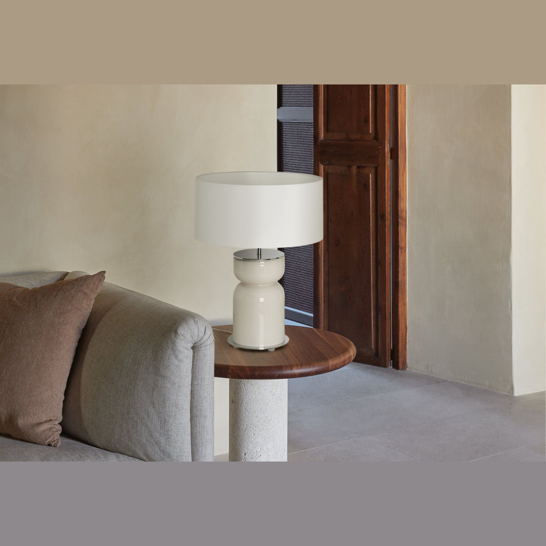 Cream glass table lamp with white shade on a wood side table beside a grey sofa