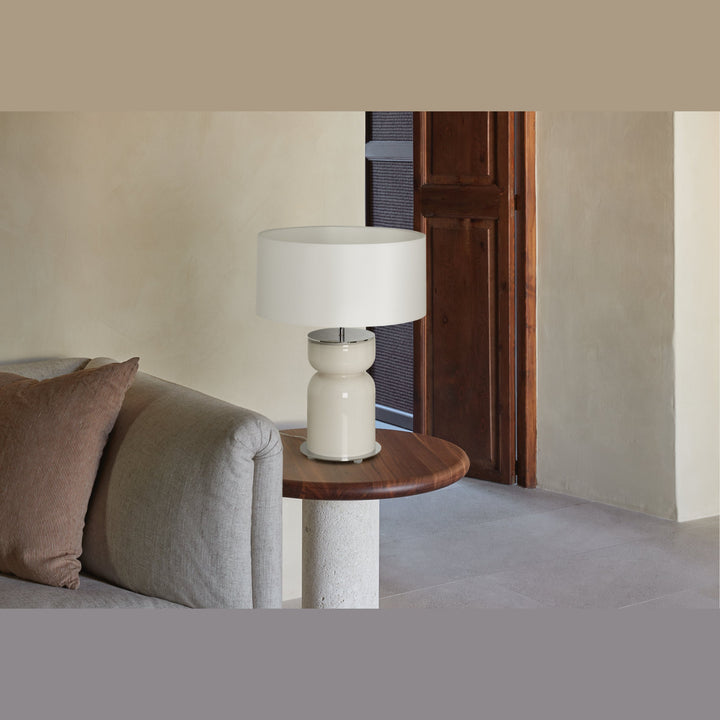 Cream glass table lamp with white shade on a wood side table beside a grey sofa