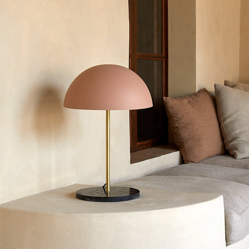 Hedra Table Lamp with a brass stem and blush dome shade on a plaster bench beside a cushioned sofa