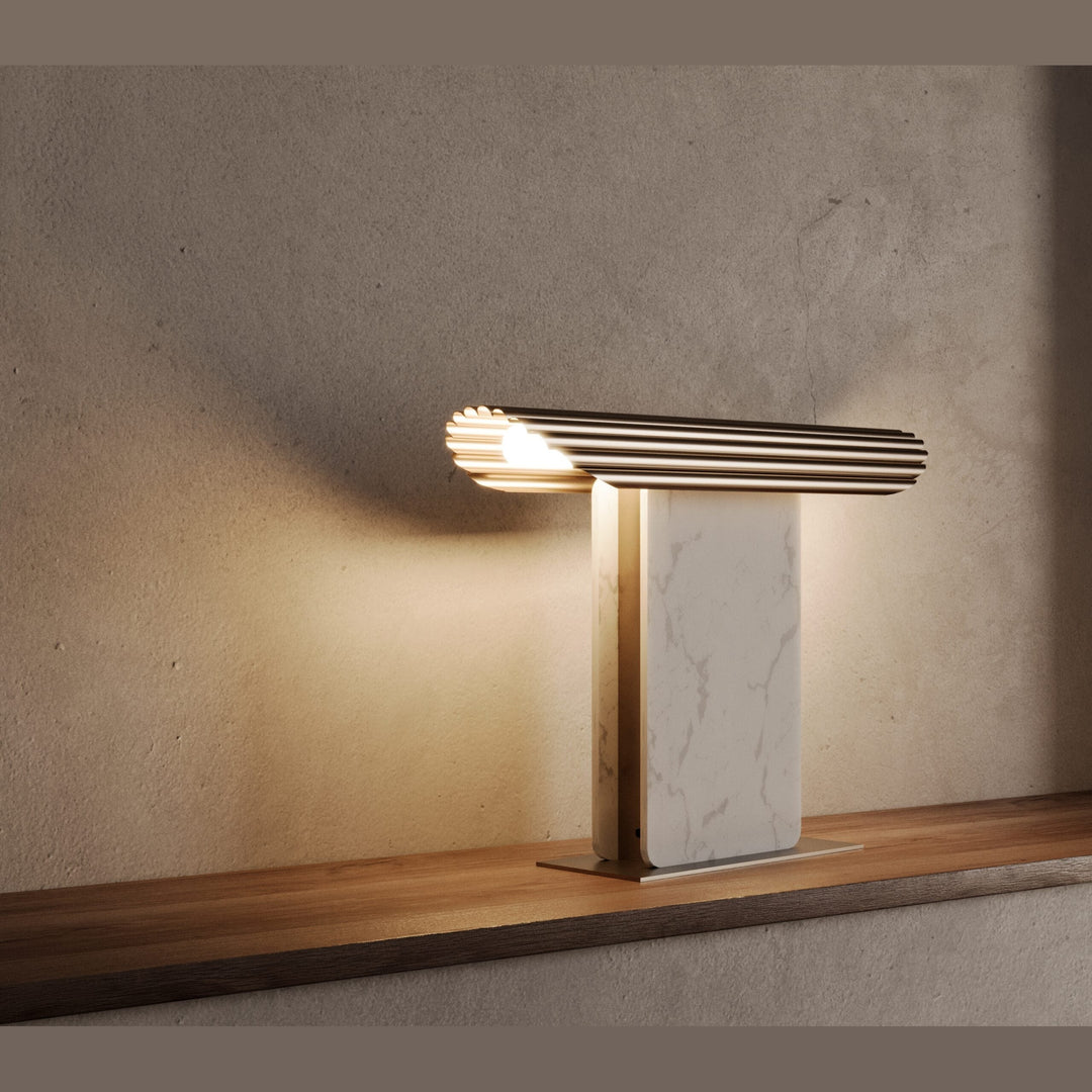Lit white marble table lamp with ribbed gold top glowing on a wood shelf against a textured wall