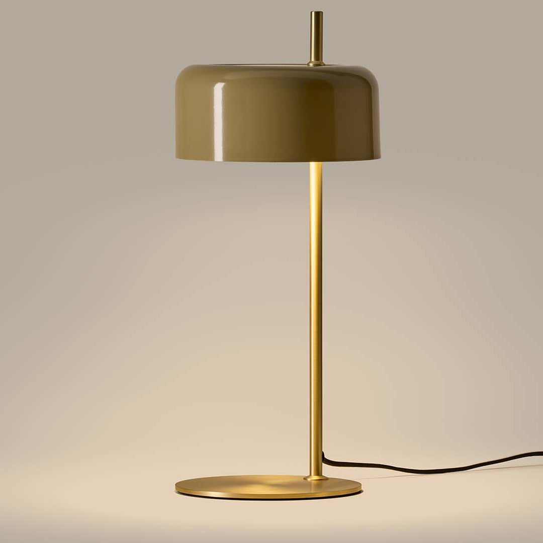 Glossy olive ceramic shade on a slim brass stem and round base, lit softly against a beige wall