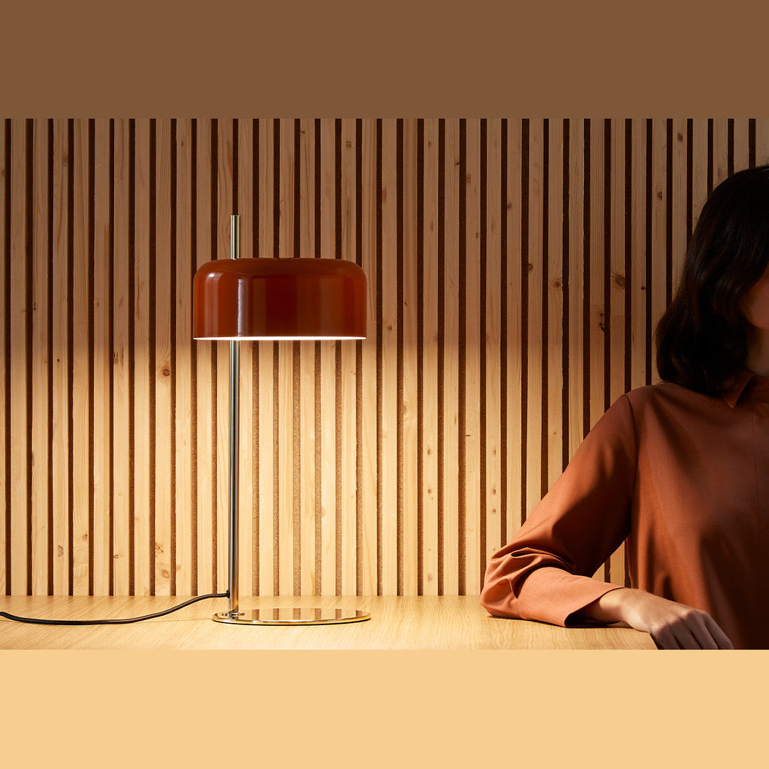 Red ceramic shade with brass stem glowing on a table in front of slatted wood panels, woman seated beside