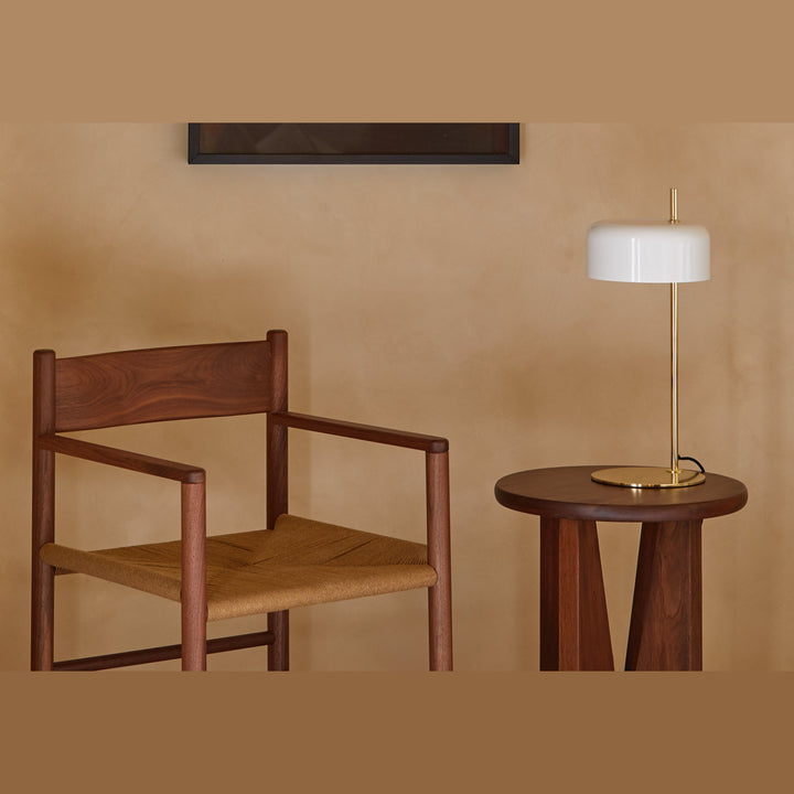 White-shade lamp lit on a dark wood side table next to a rush-seat wooden armchair