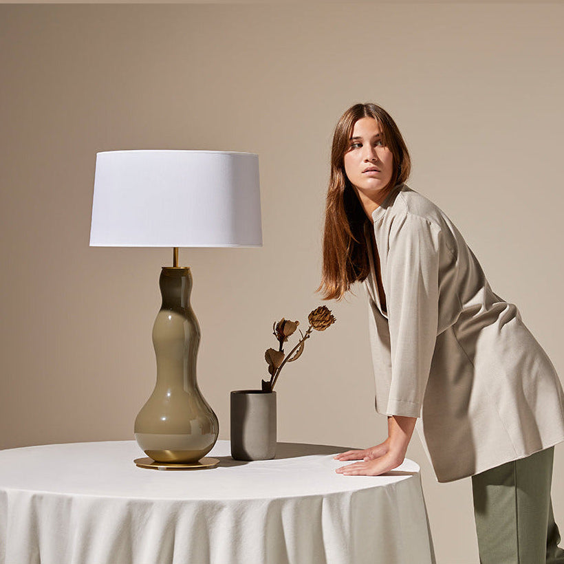 Melly Table Lamp on a round linen-draped table next to a woman and a vase of dried flowers