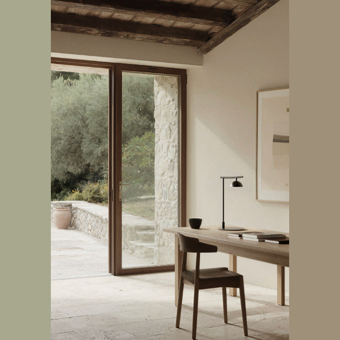 Tana Table Lamp in black on a wooden desk by a glass door overlooking an olive garden