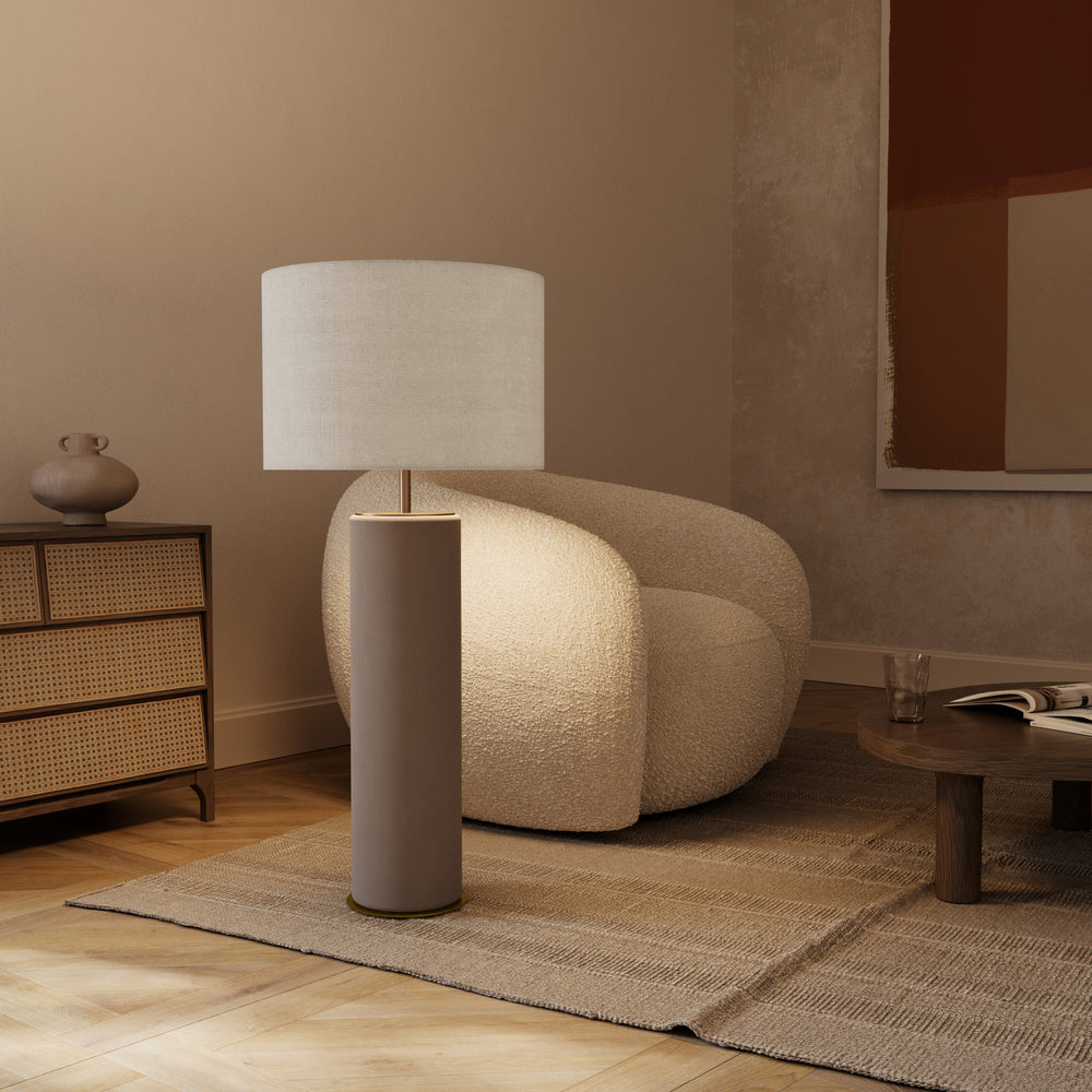 Amec Table Lamp in taupe ceramic standing on a woven rug beside a boucle armchair and cane dresser, lit