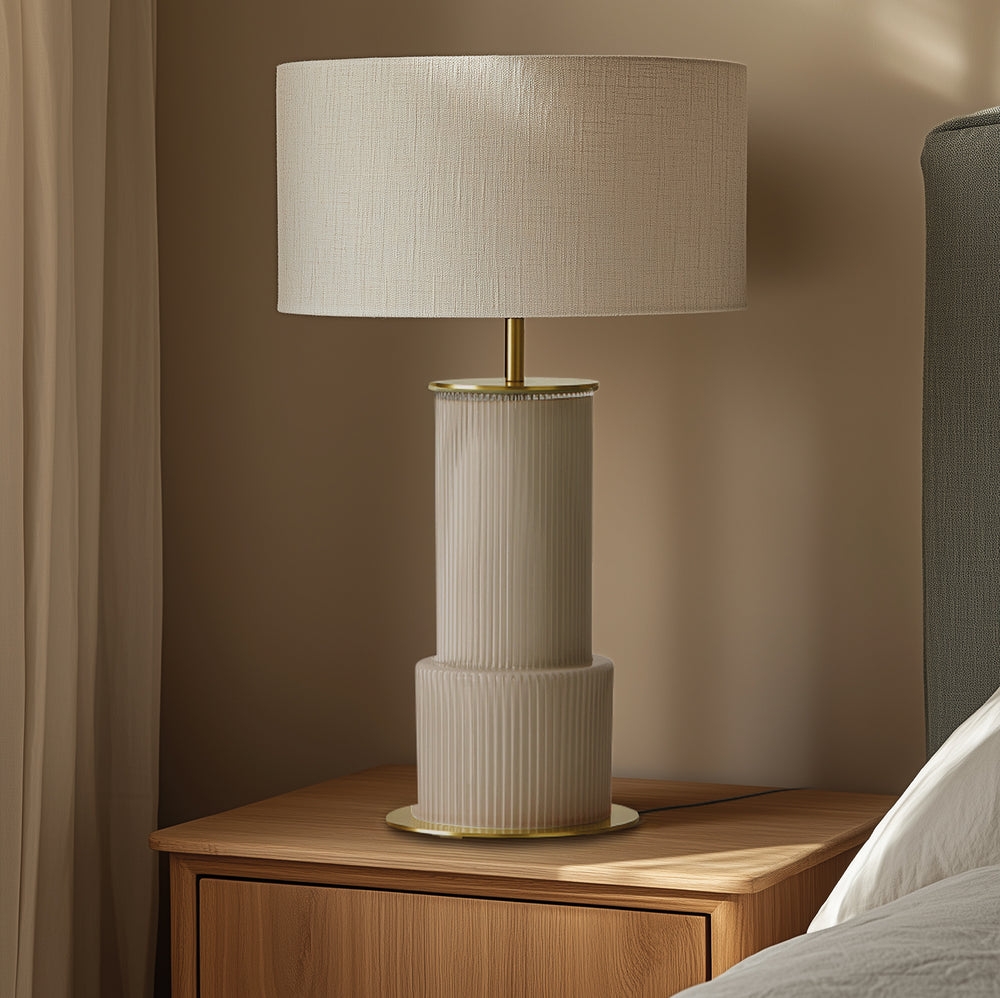 Atina Table Lamp with linen shade and brass stem on a wooden nightstand beside a bed, lit