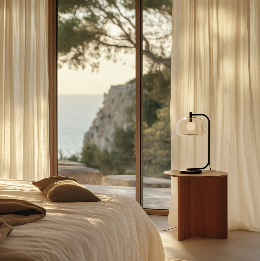 Cloud Table Lamp with clear glass shade on a wooden bedside table by a bed, sheer curtains and sea view