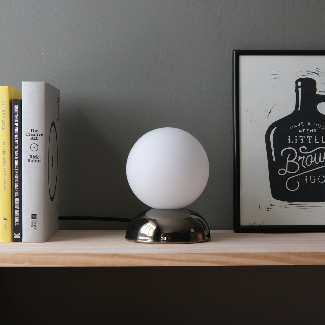 Sleek white globe lamp with chrome base, styled beside books and framed art on a wooden shelf.