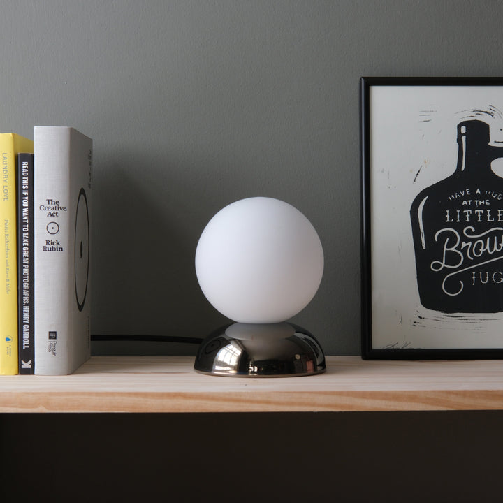 Sleek white globe lamp with chrome base, styled beside books and framed art on a wooden shelf.