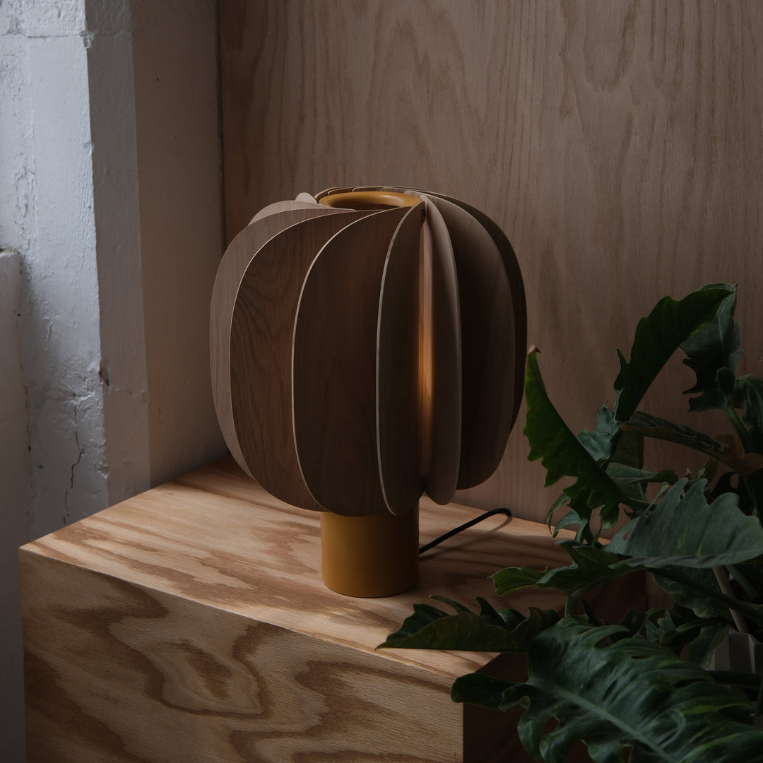 Mid-century modern wooden table lamp with sculptural petal design, warm amber base, styled with lush greenery on oak surface