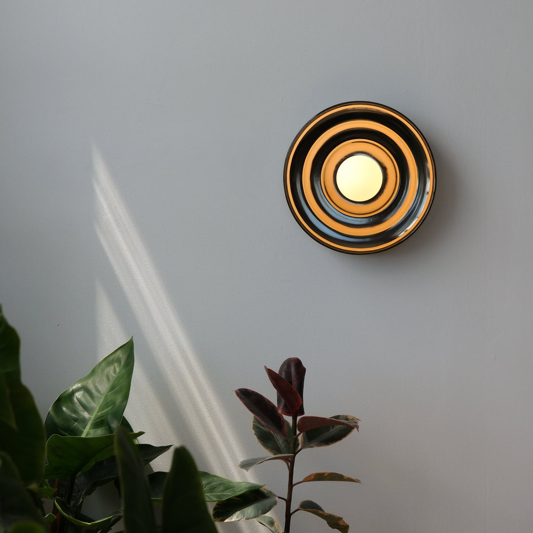 Circular brass wall sconce with concentric rings, casting warm glow beside lush greenery