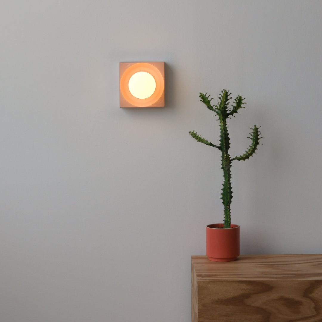 Square ceramic wall sconce with warm glow beside potted cactus on oak console