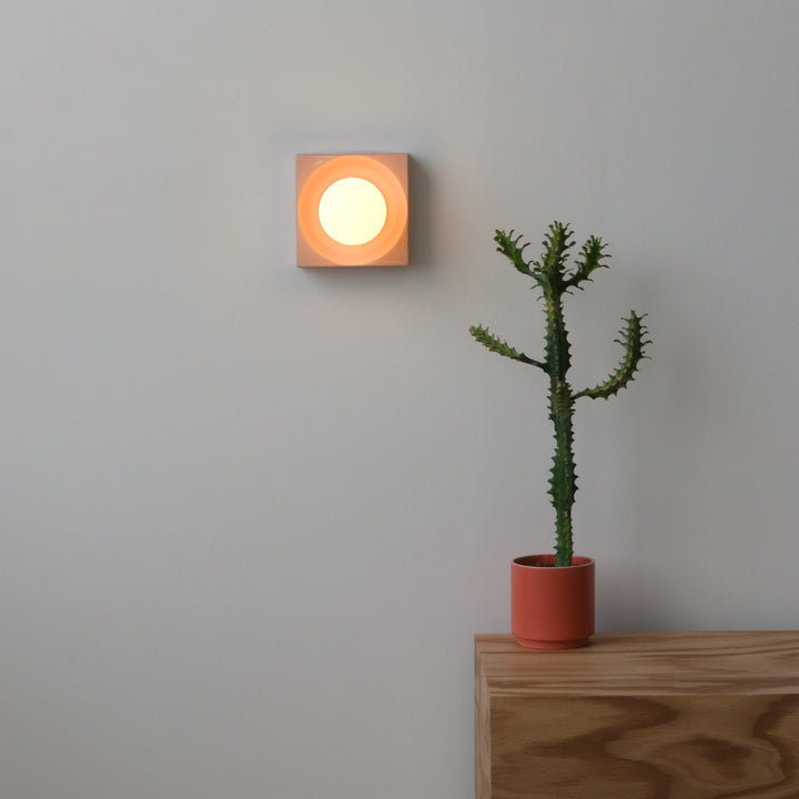 Square ceramic wall sconce with warm glow beside potted cactus on oak console