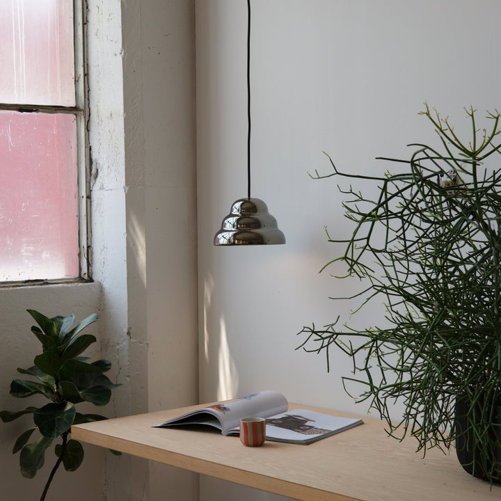 Chrome pendant light above wooden desk with plants and open magazine, minimalist home decor