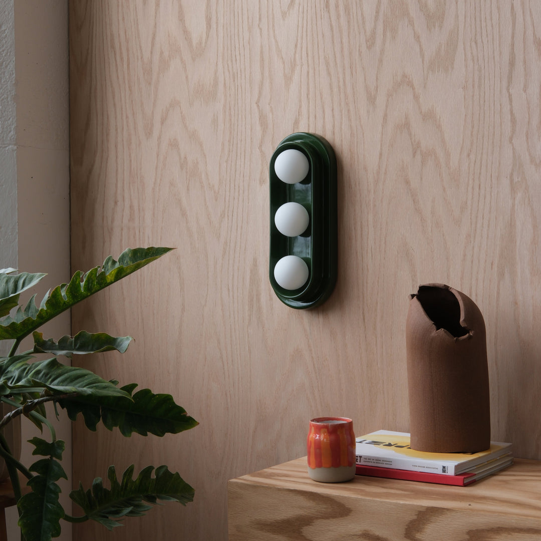 Emerald green wall sconce with white globes, styled with ceramic vase and books on wood shelf