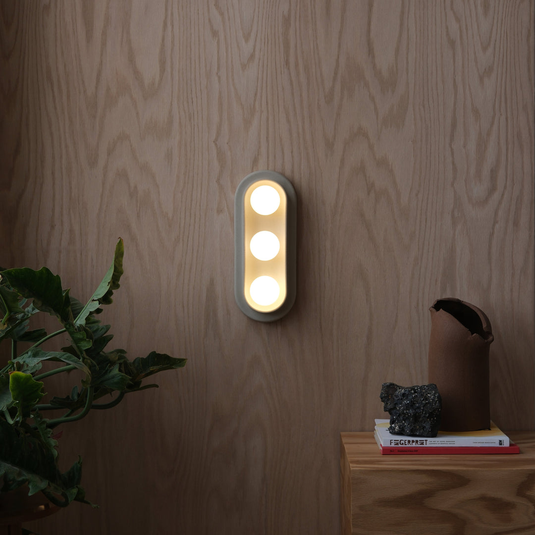 Modern wall sconce with three circular lights, mounted on wood-paneled wall, styled with greenery and decor