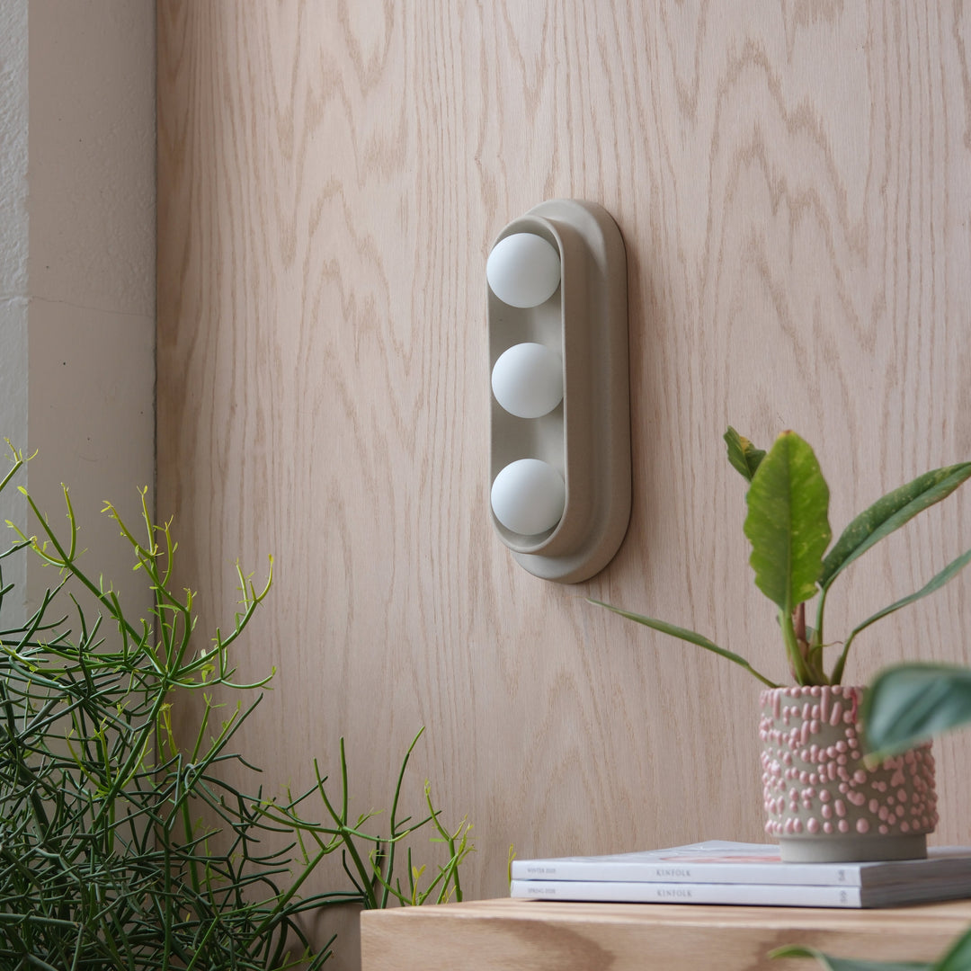 Modern wall sconce with three frosted glass globes, mounted on light wood paneling, styled with greenery and books.