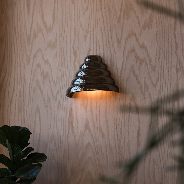 Glossy ceramic wall sconce with tiered design, casting warm glow against wood paneling, styled with fiddle leaf fig.