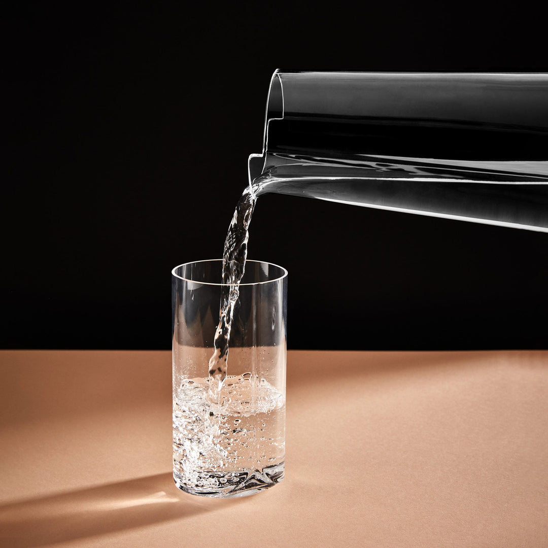 Hew Highball Glass, Set of 4 — water poured from a carafe into a clear highball glass on a tan surface