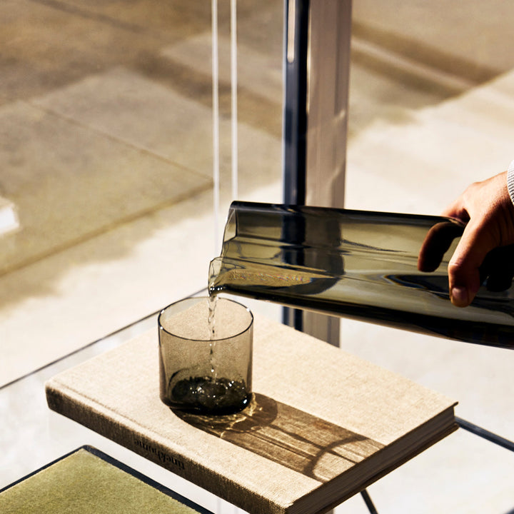 Hew Tumbler, Set of 4 — water poured from a Smoke carafe into a Smoke tumbler on a book