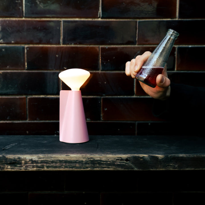 Pink ceramic table lamp with warm glow beside hand holding glass bottle against brick wall