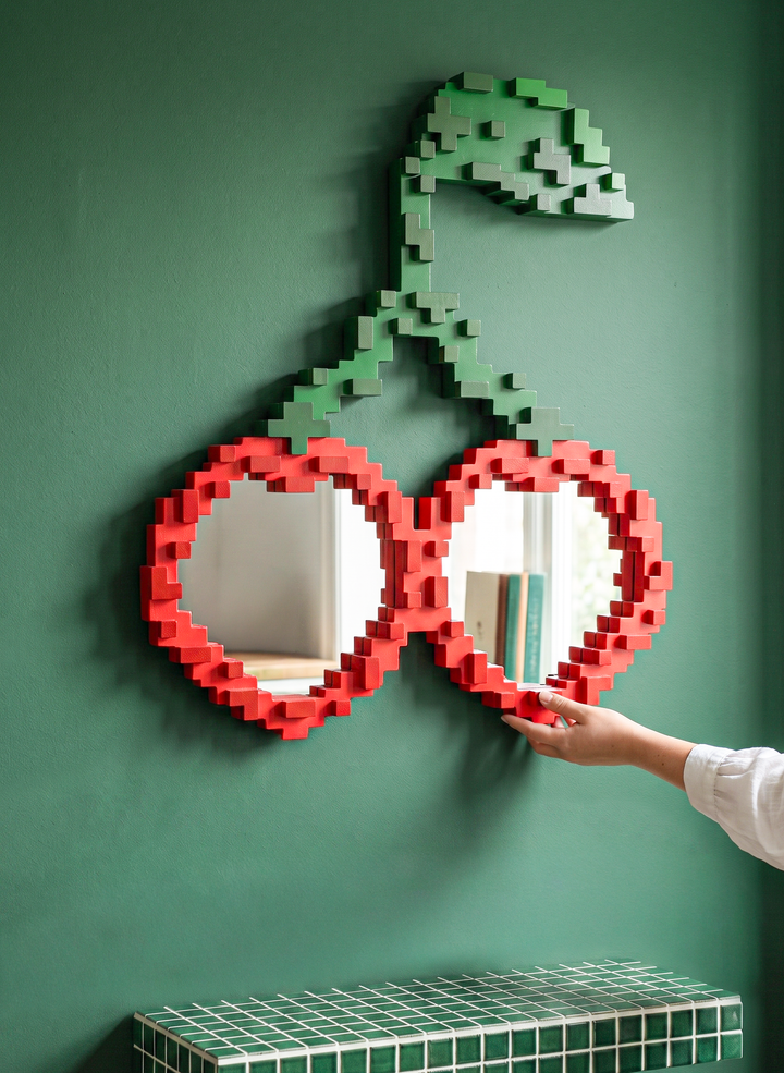 Seletti Pixel Mirror Cherries, the cherry mirror hung on a green wall above a grid console, a hand touching one lens