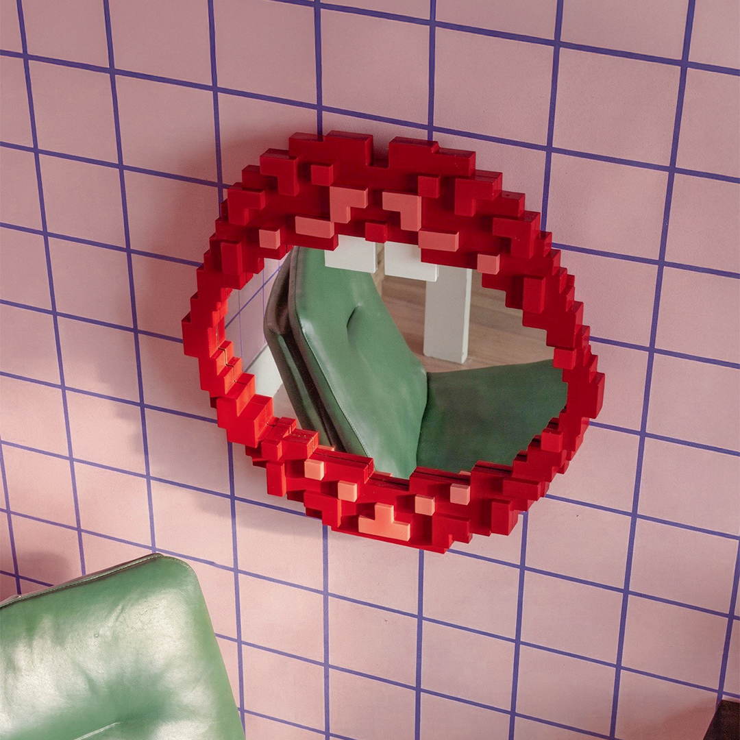 Seletti Pixel Mirror Mouth, the mouth mirror on a pink grid-tiled wall above a green cushion