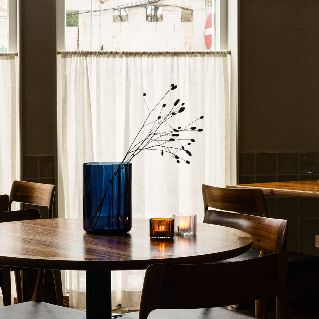 Pulse Tealight Holder X — tealight holders lit on a wooden cafe table beside a dark blue vase with branches