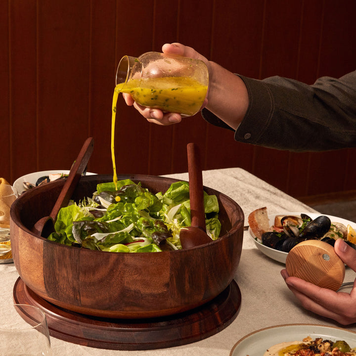 Person pouring dressing over a salad in a wooden bowl on a dining table.
