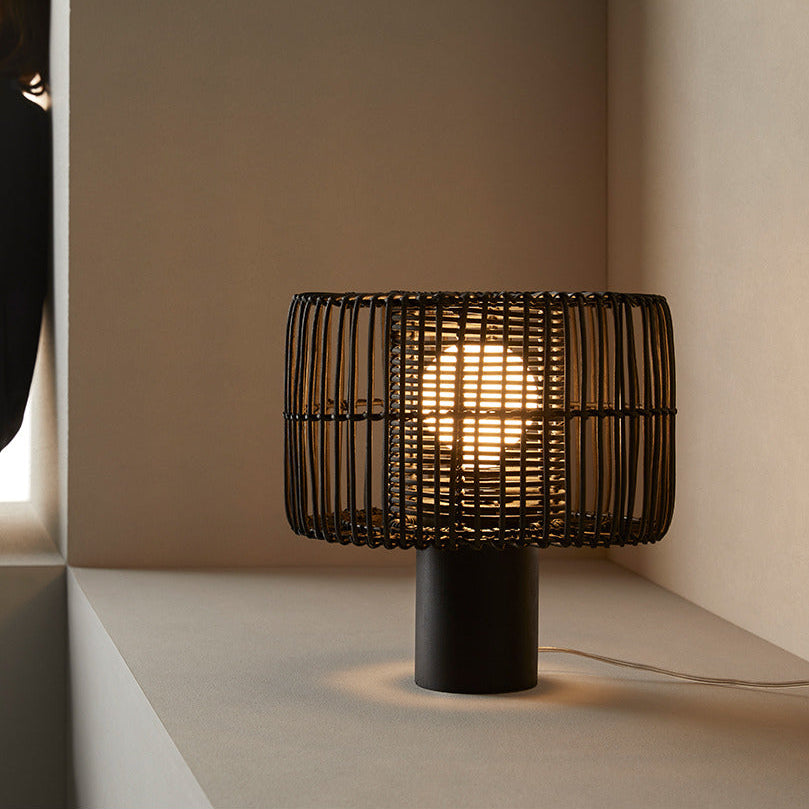 Hedula Table Lamp lit on a stone ledge, its black rattan shade glowing, with a woman in black seated nearby