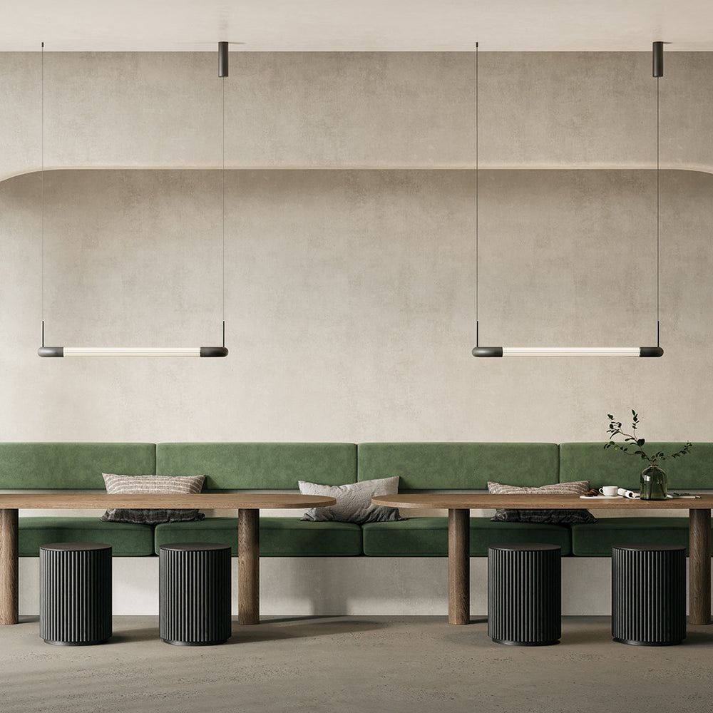Two Iman Linear Pendants lit above long wooden tables and a green banquette in a neutral dining space
