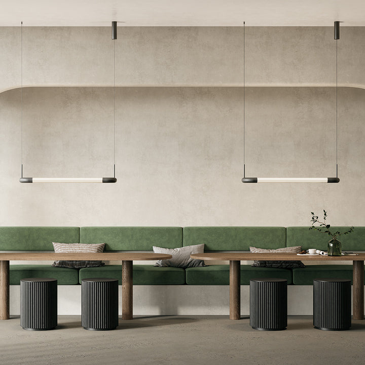 Two Iman Linear Pendants lit above long wooden tables and a green banquette in a neutral dining space