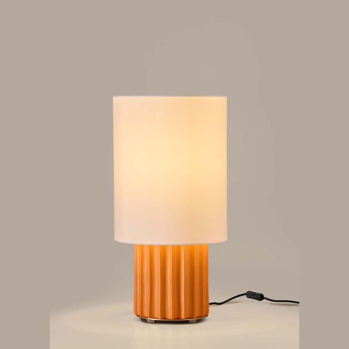 Lit table lamp with orange fluted ceramic base and cylindrical white shade on a grey backdrop