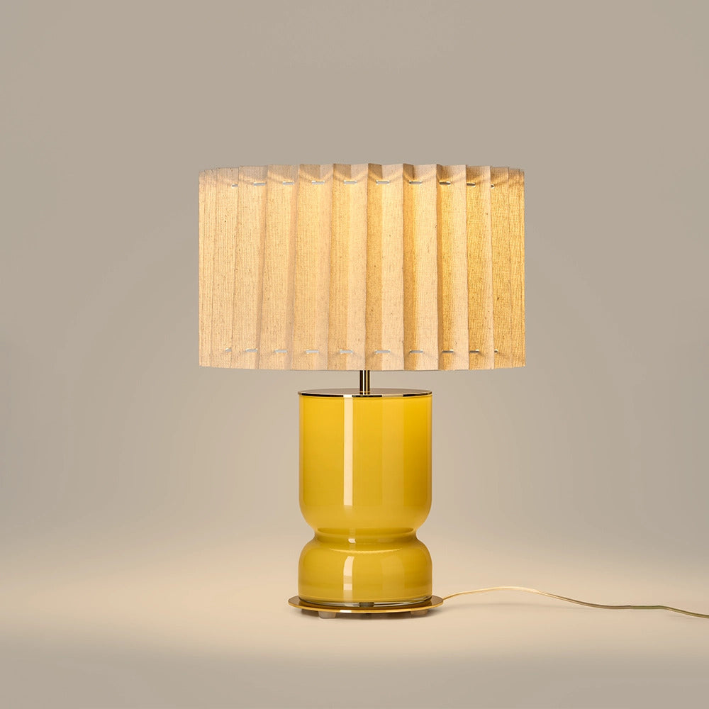 Lit table lamp with glossy mustard yellow glass base and pleated linen shade