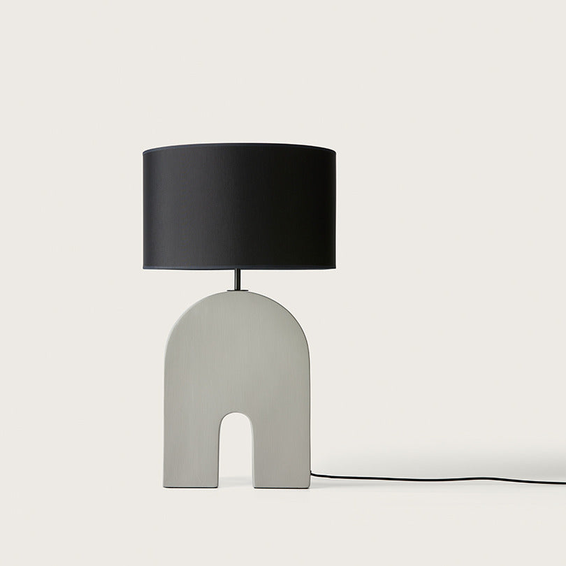 Home Table Lamp in Ash Grey with an arched ceramic base and black drum shade, unlit, by Aromas del Campo